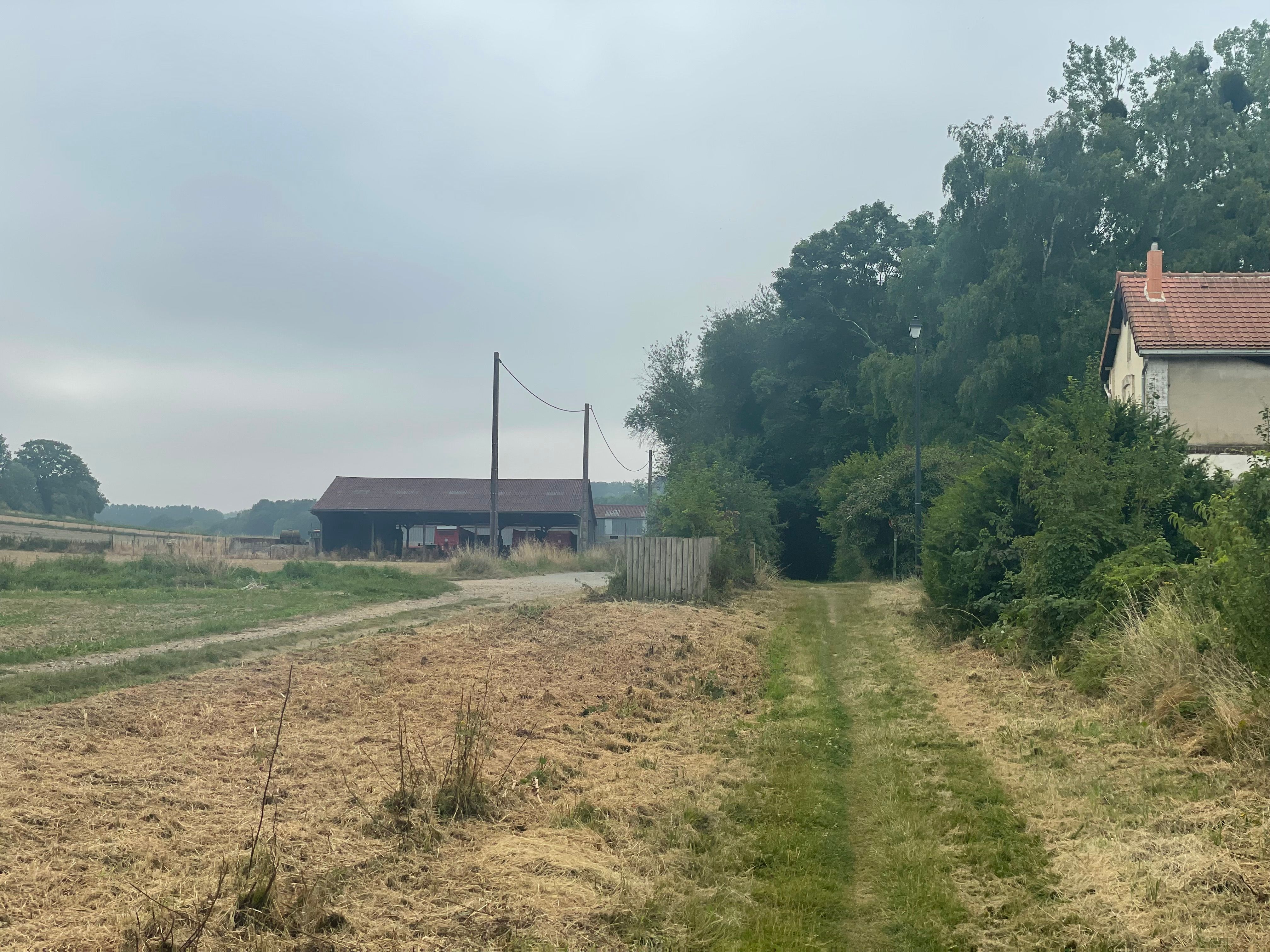 La voie verte, dans un des rares endroits dégagé. On distingue à droite un ancien poste de passage à niveau, et à gauche, un peu plus loin, un hangar de ferme. Le ciel est gris, voilé