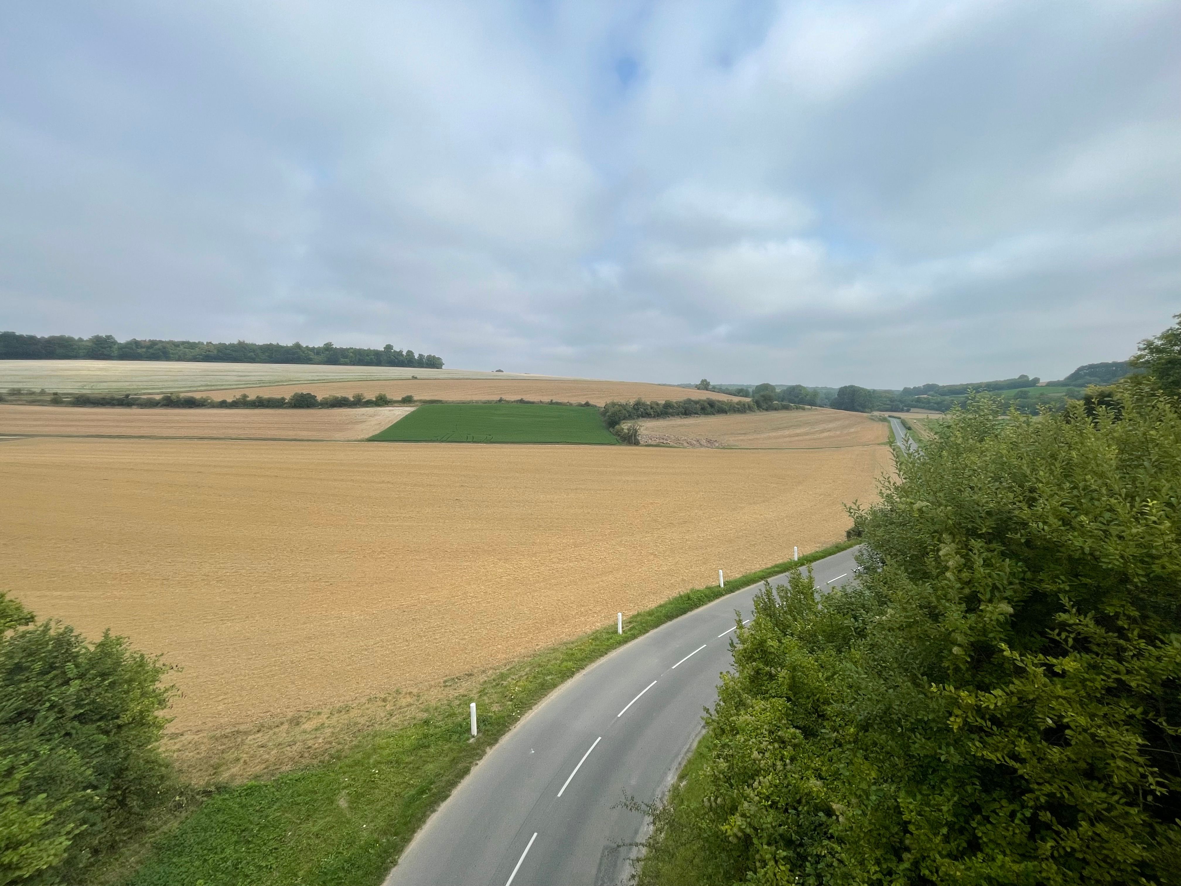Paysage de campagne pris au grand angle. Une route tourne et s'échappe du cadre par la droite. Au fond, les champs semblent dessiner un motif géométrique
