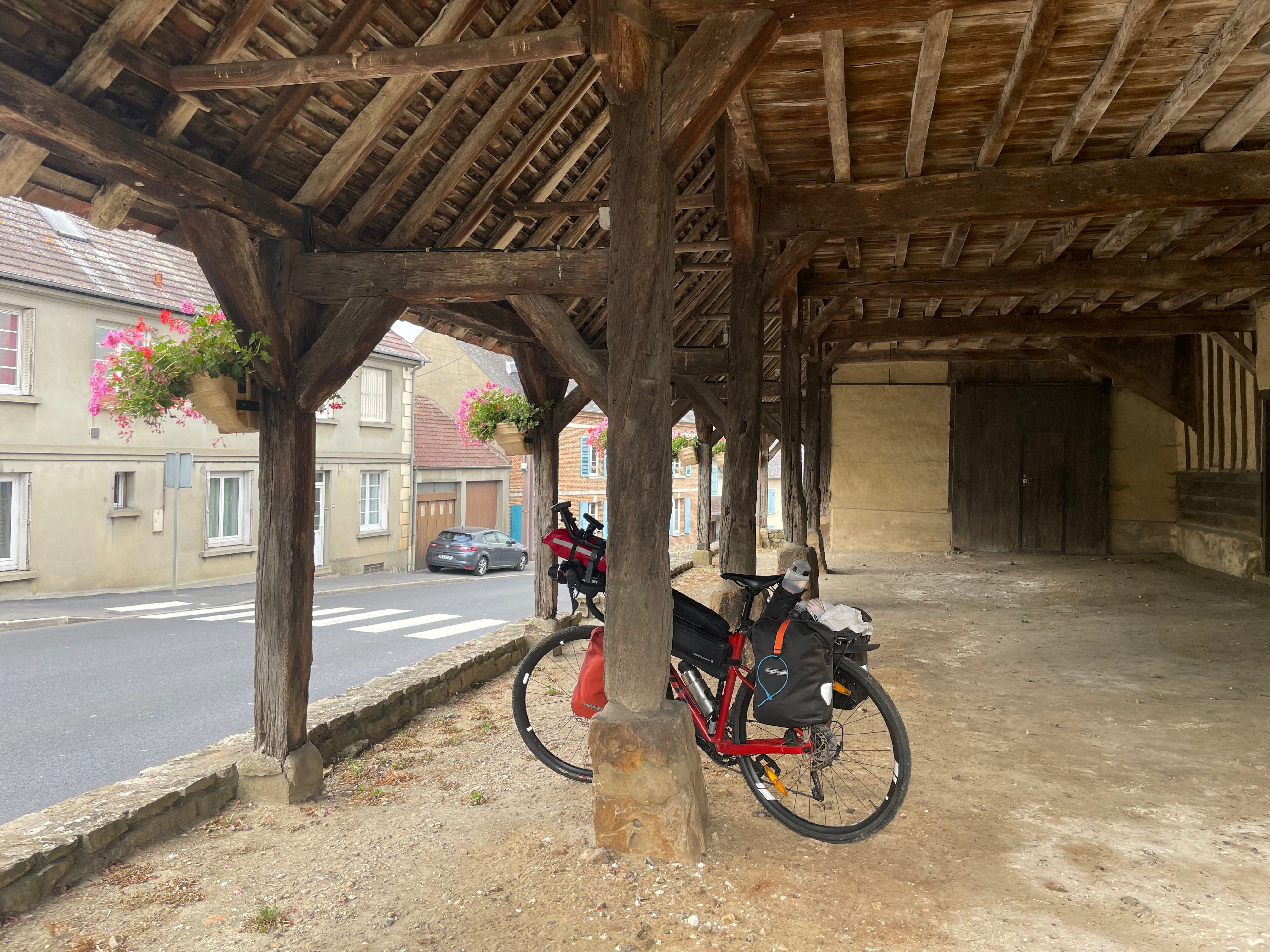 Photo du vélo, adossé au pilier en bois d'une halle couverte. La charpente de celle-ci est visible.