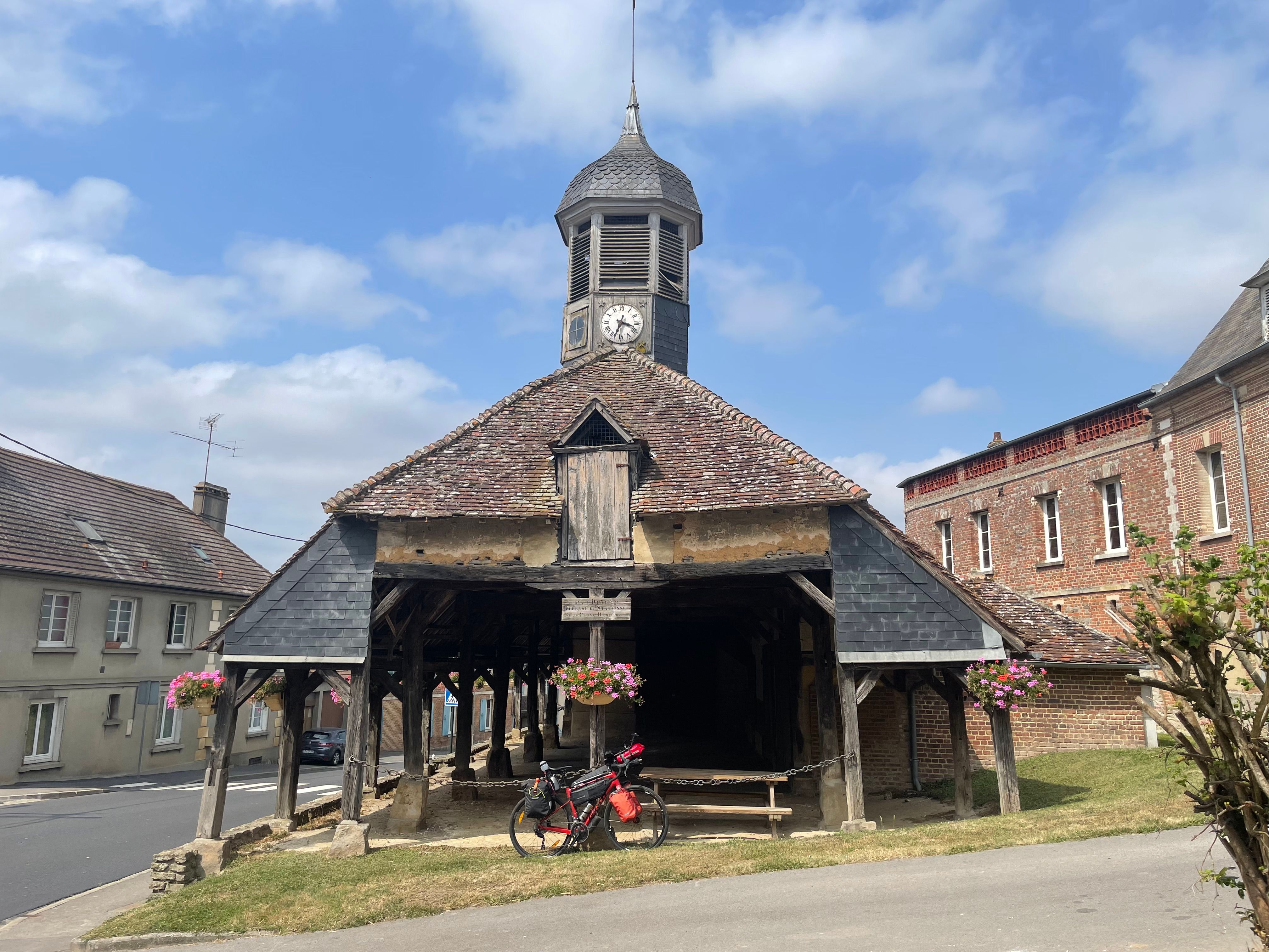 Vue d'extérieur de la halle couverte. Le toit est récouvert de tuiles et d'ardoises. Une genre de petit clocheton abrite une horloge HS, ainsi qu'un carillon