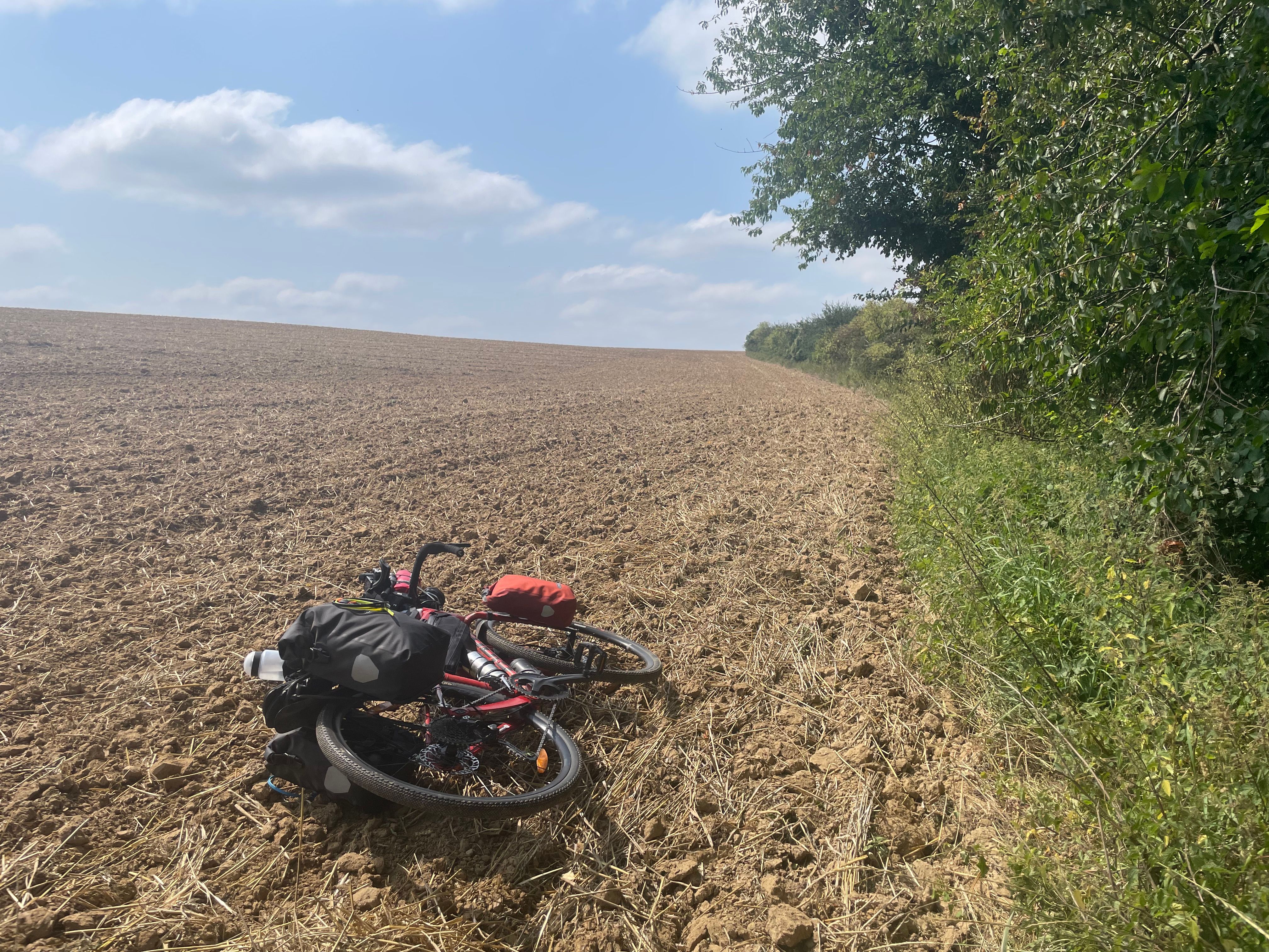 Un vélo rouge avec ses sacoches est couché sur le flanc dans un champ déchaumé. Le champ s'étend vers l'horizon, et une haie le borde sur la droite. Le temps est ensoleillé et le ciel bleu, avec quelques petits nuages