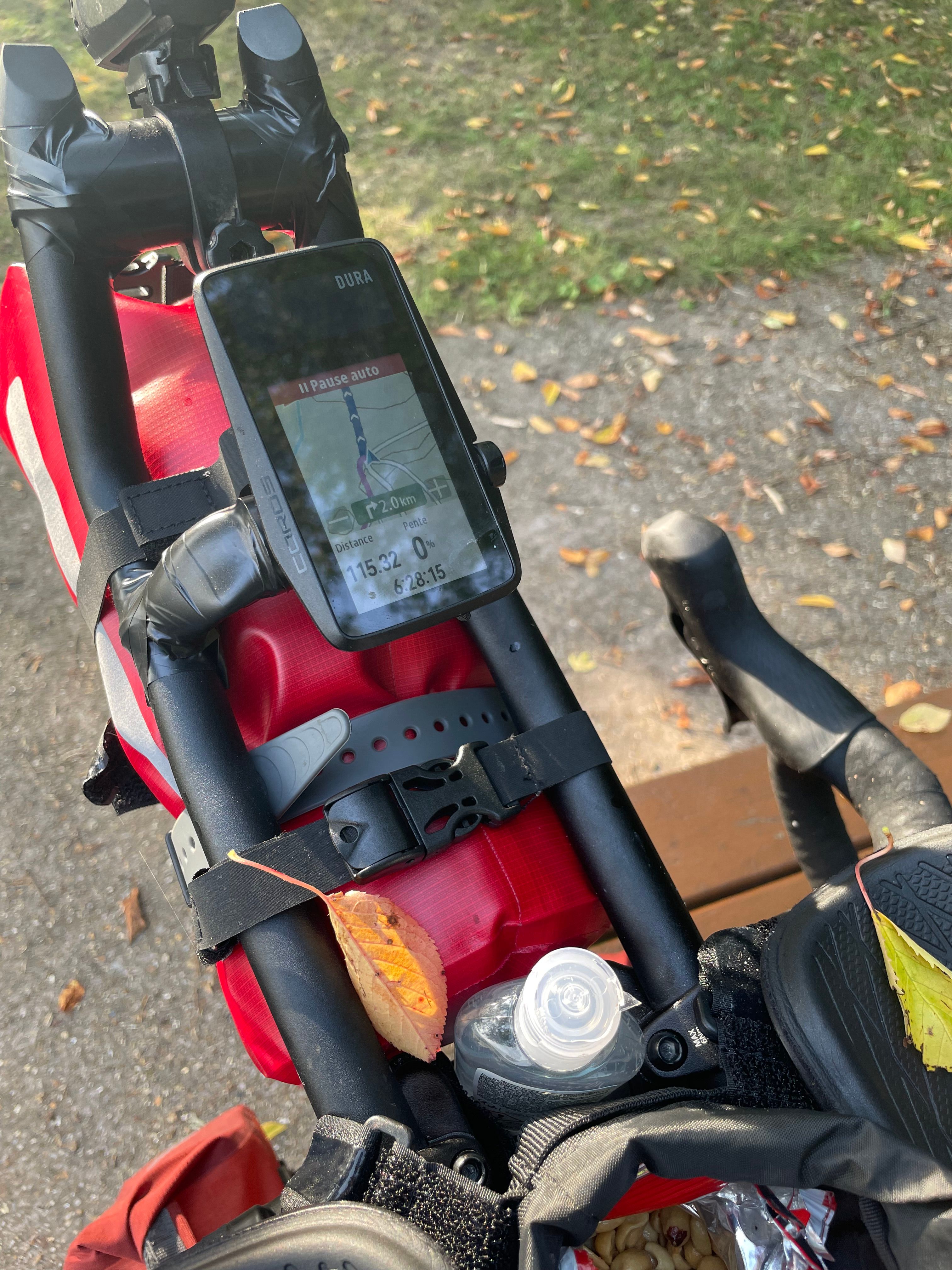 Vue du cockpit du vélo. Le GPS du vélo est fixé sur les prolongateurs. On peut y lire 115.22km. Des feuilles mortes se sont posées sur le sac rouge fixé lui aussi sur les prolongateur