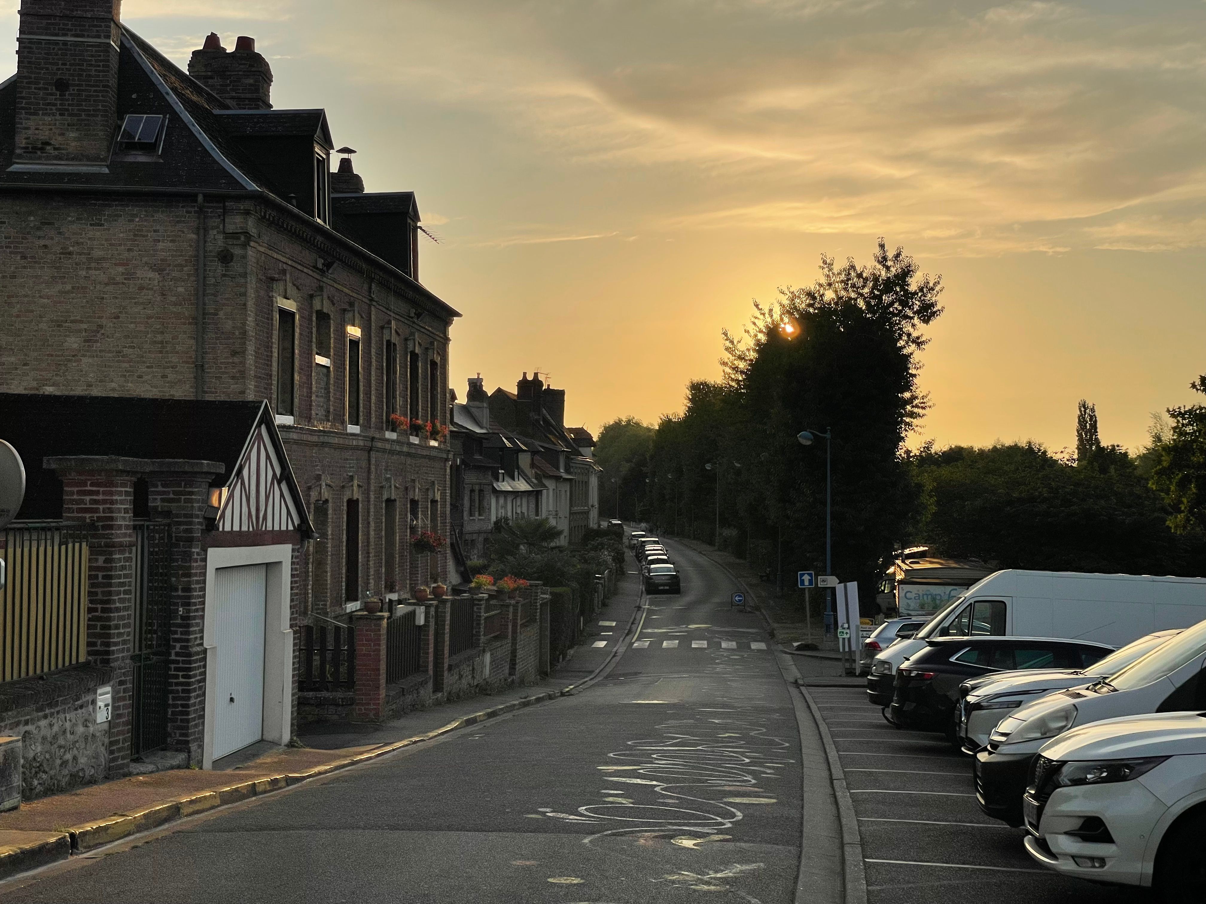Vue au soleil couchant d'une rue de Pont-de-l'Arche longeant la Seine et l'Eure. Le soleil est caché par un arbre et le ciel a un très beau dégradé dans les teintes dorées