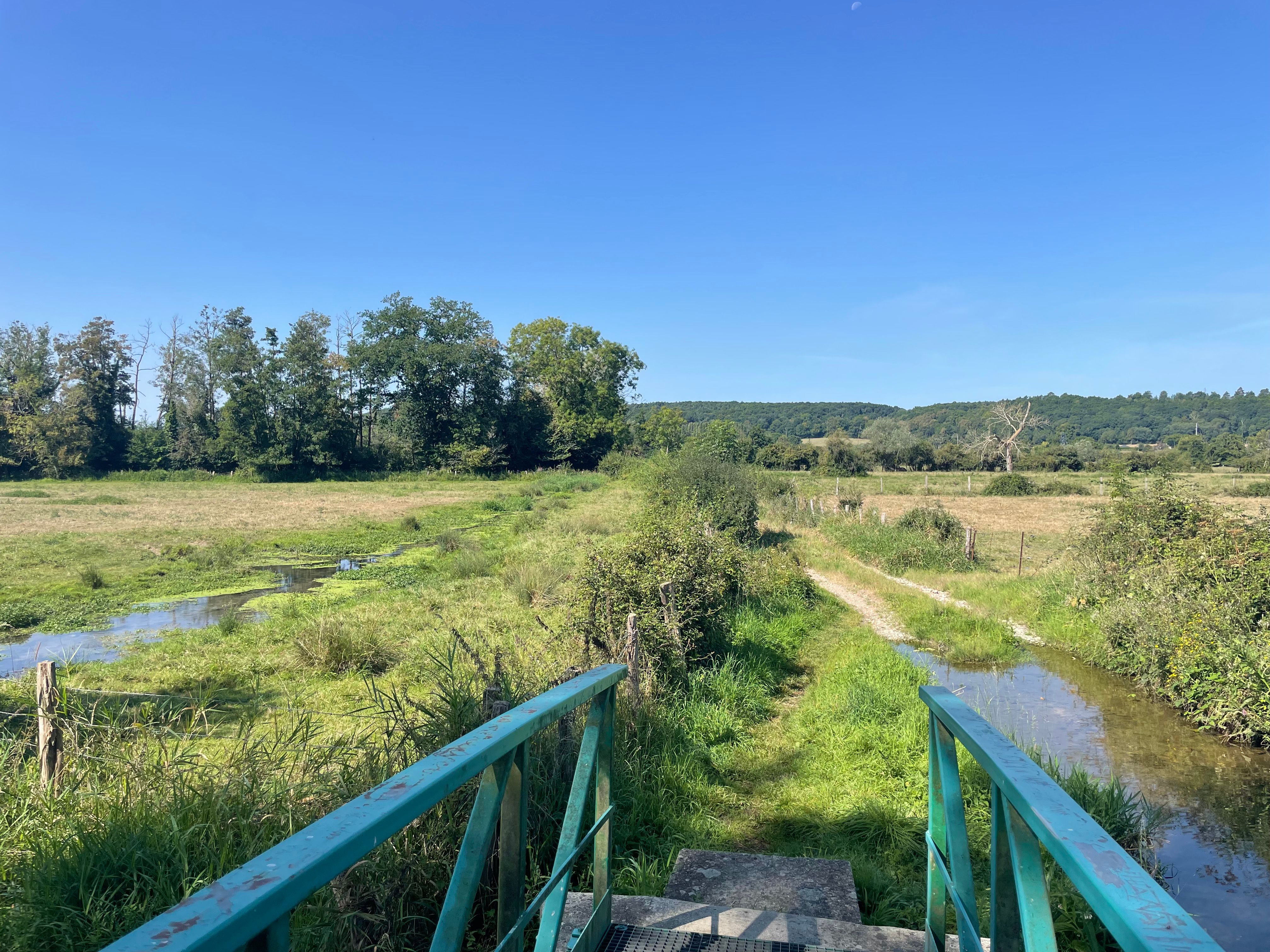 Vue sur la campagne normande depuis un petit pont (dont on distingue les rambardes vertes). Sur la droite, le chemin inondé se transforme en gué. En arrière plan, des haies et 2 collines au loin