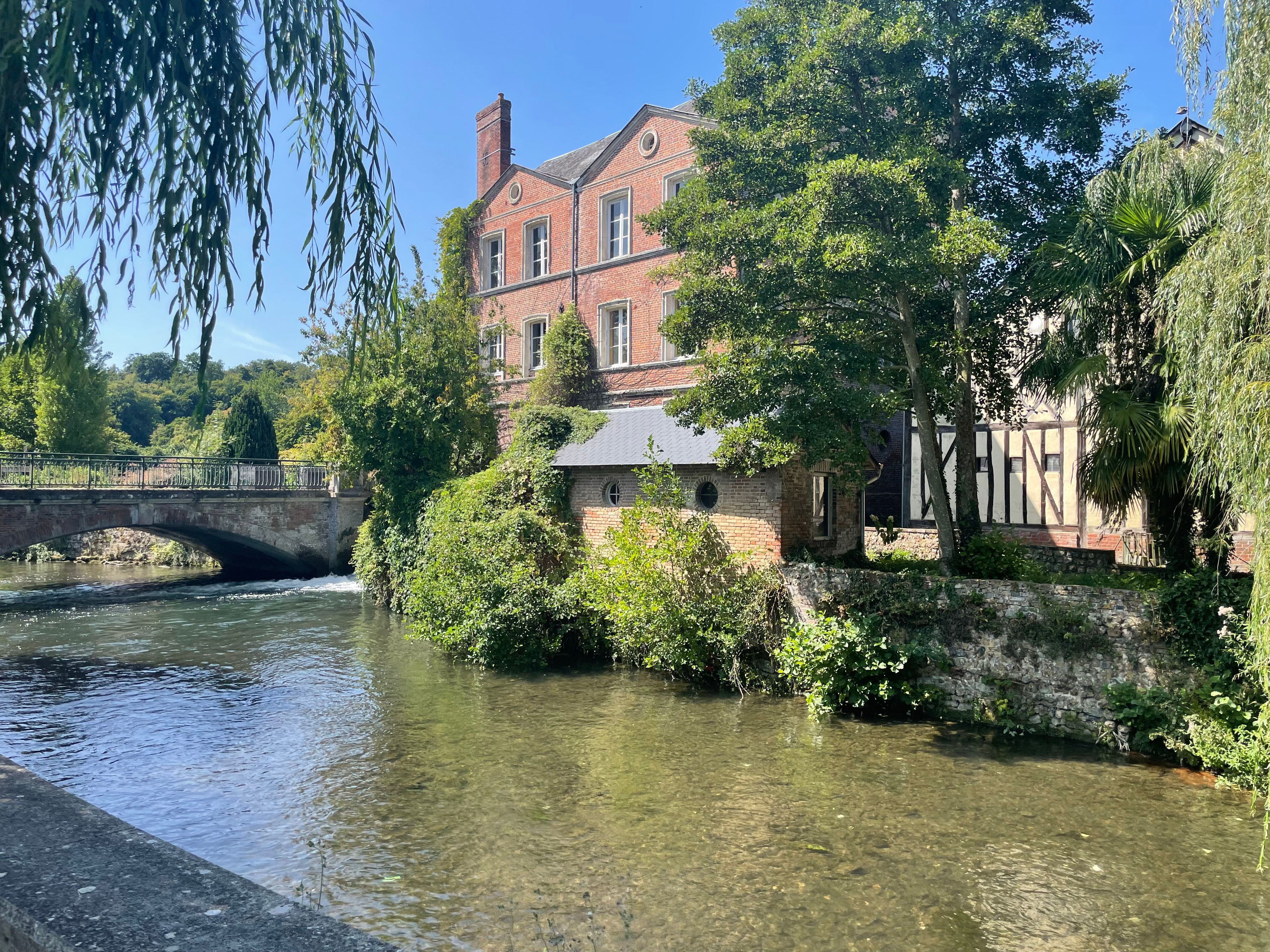 Vue sur l'ancien moulin de Broglie et le pont au dessus de la Charentonne. La lumière est forte. Le pied des murs du moulin est envahi de végétation qui a les pieds dans l'eau