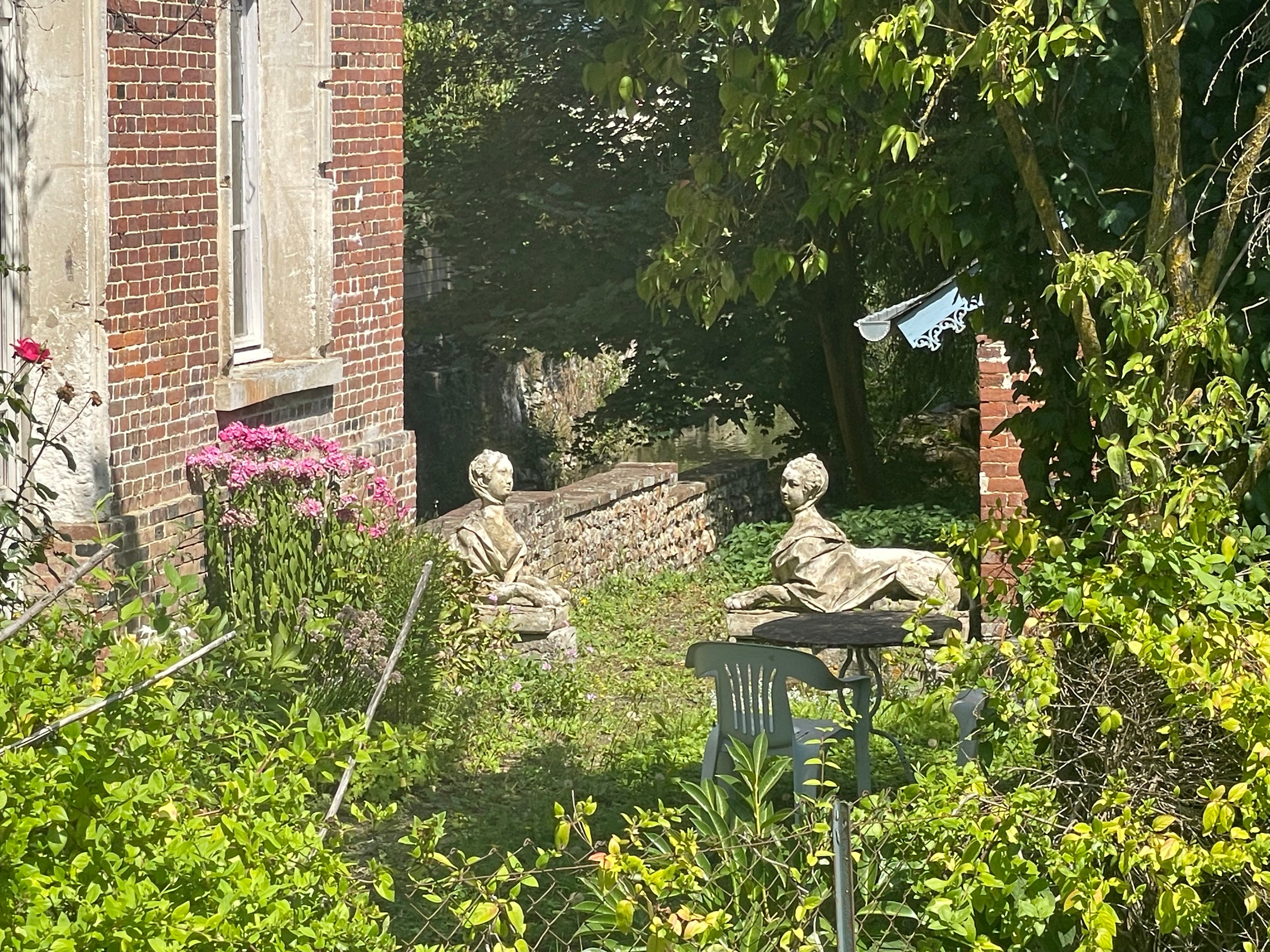 Deux statues de Sphinx se font face dans un petit jardinet envahi de végétation. On distingue une chaise et une table de jardin devant le sphinx de droite