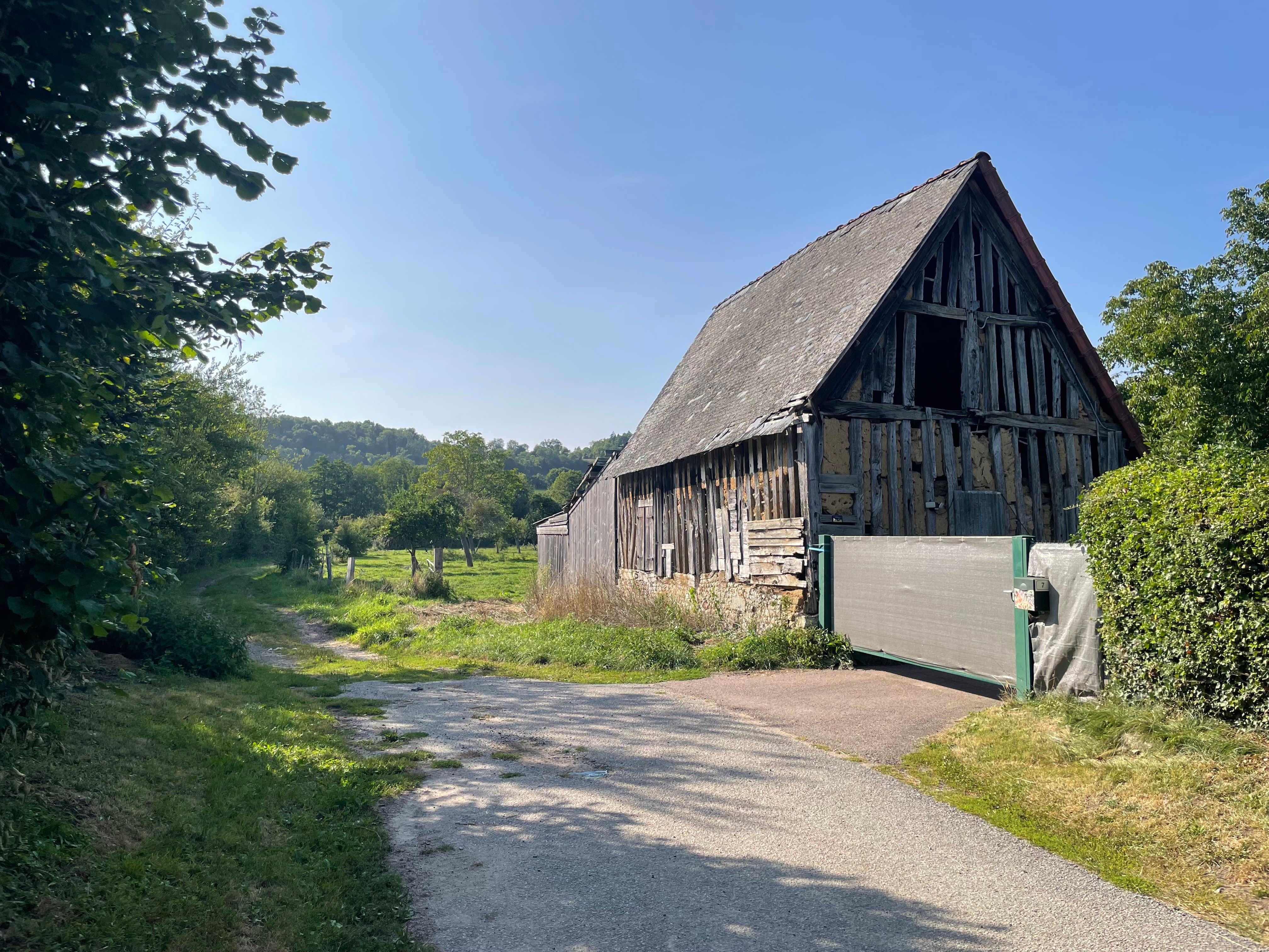 Debut d'un chemin grimpant une colline verte. Le chemin est entouré de haie, et au premier plan, l'entrée de celui-ci est bordé d'une vieille grange en torchis et colombages, flanquée d'un portail vert moderne