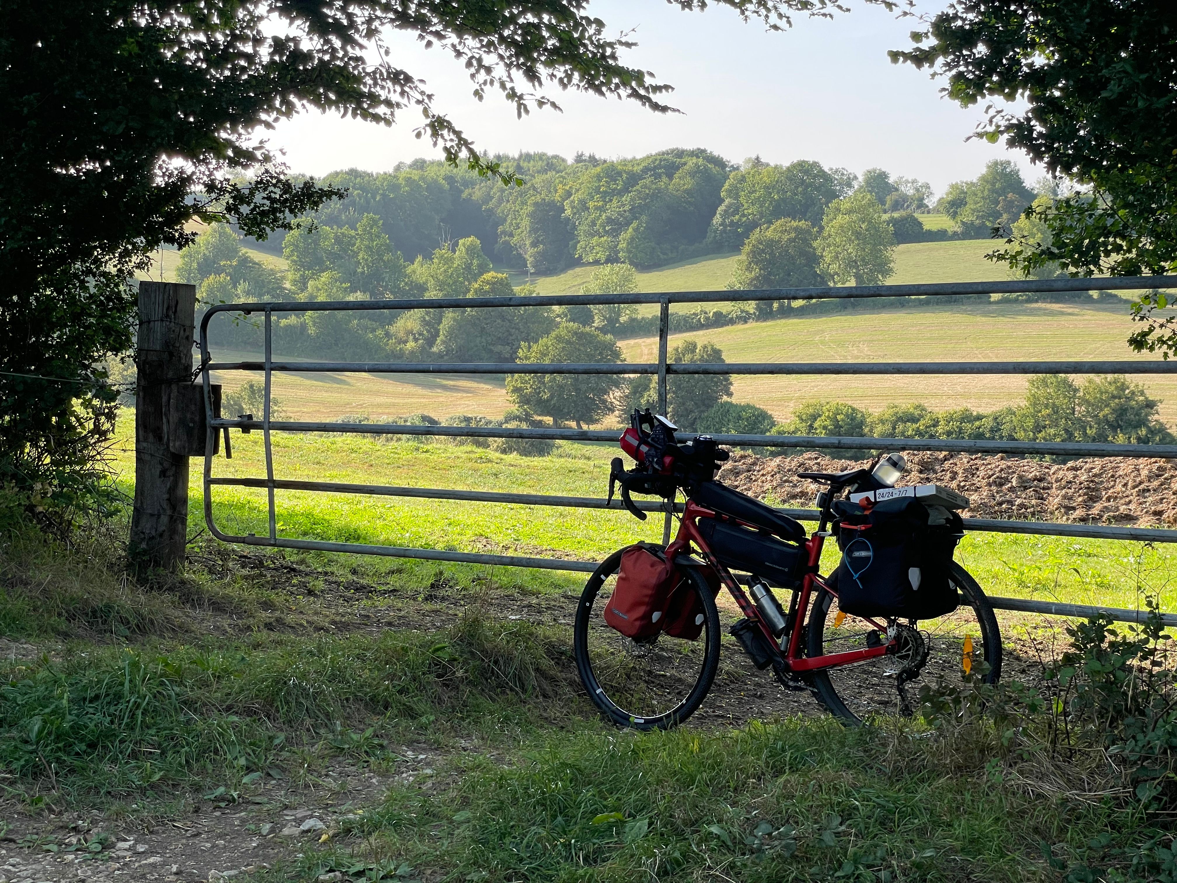Le vélo, chargé de ses sacoches et d'un carton de pizza est posé sur la barrière d'une pature. Le feuillage des arbres environnants dessine une arche par dessus. Derrière la barrière on voit une pature en pente, baignée de lumière de fin d'après-midi