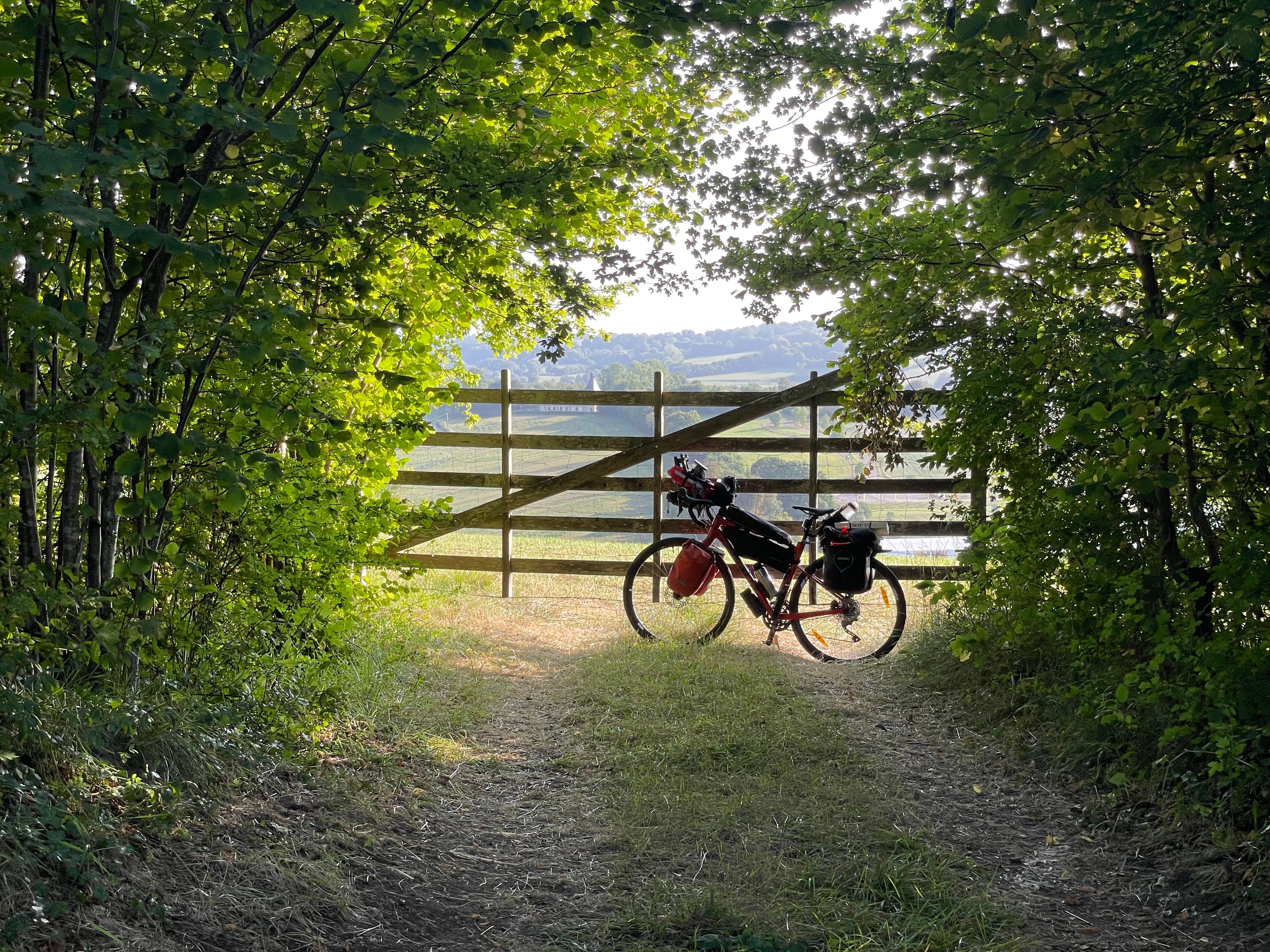 Velo posé sur une barrière à contre-jour, au bout d'un chemin. Le chemin est bordé de noisetiers, dont les feuillages proches de la barrière sont illuminés