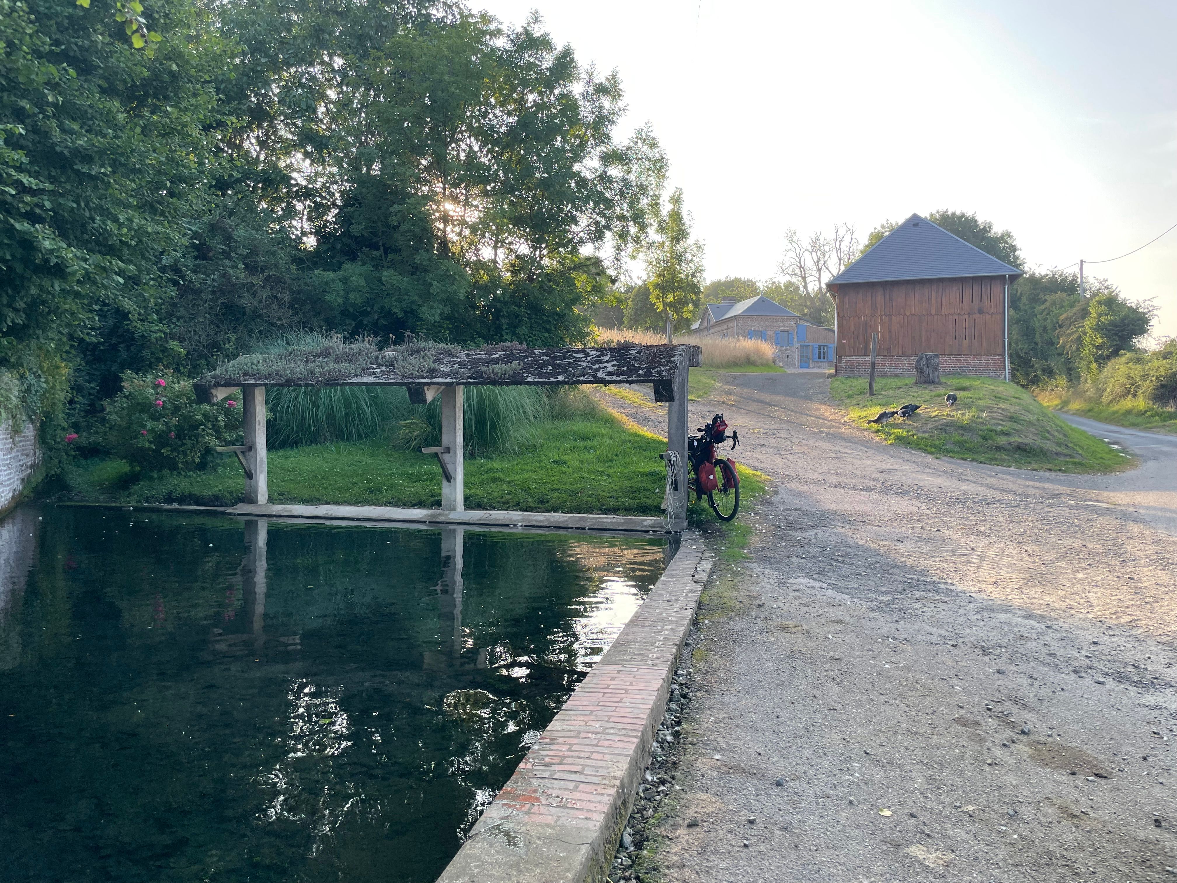 Lavoir en plein air, avec un petit auvent à son extrémité. Le vélo est adossé à l'un des piliers de l'auvent. Une petite troupe de canard noir et blanc fourrage dans l'herbe. Une ferme et une grange à l'arrière plan
