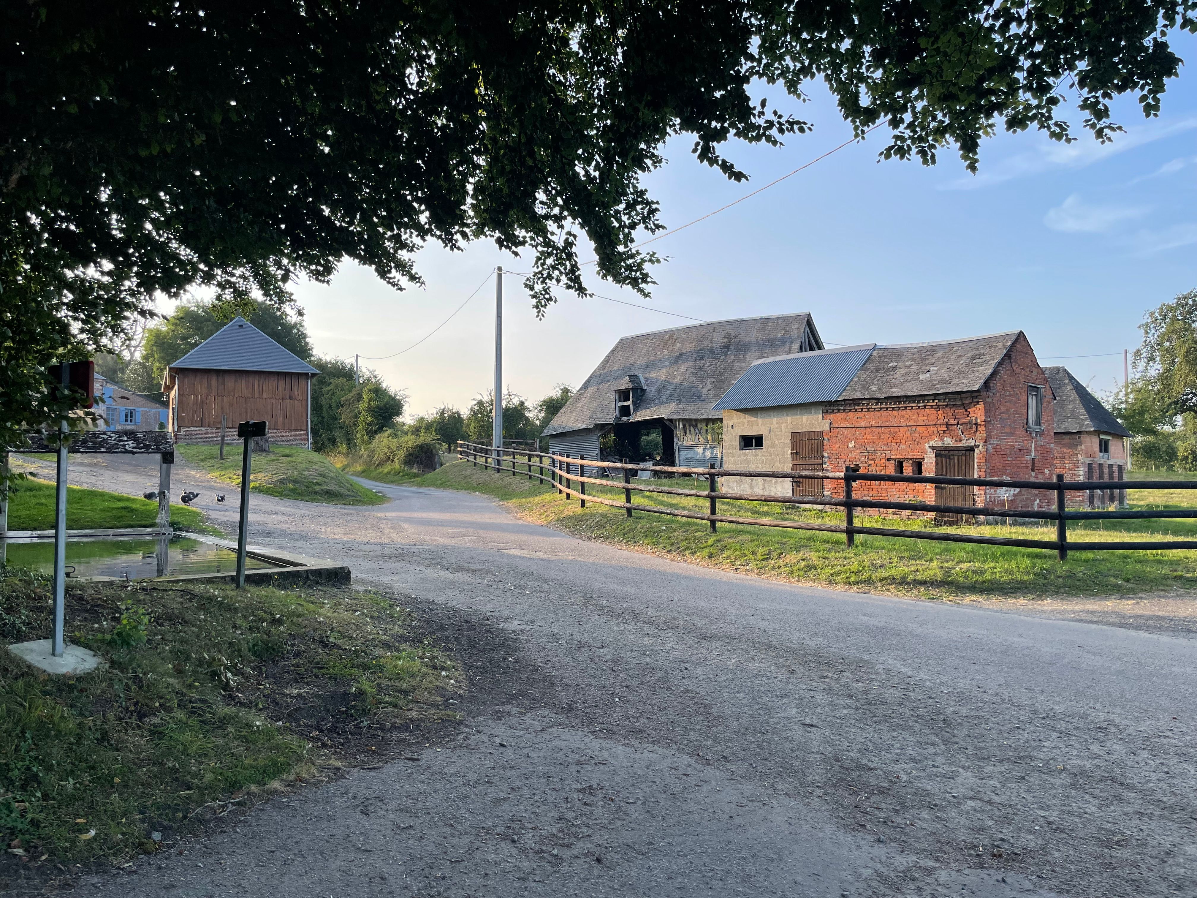 Plan large sur les batiments à proximité du lavoir. Une vieille grange et un batiment agricole retapé de bric et de broc (parpaings, briques, tôle ondulée) se trouvent derrière une cloture formée de tronc d'arbres. À l'arrière plan une route serpente en montant.