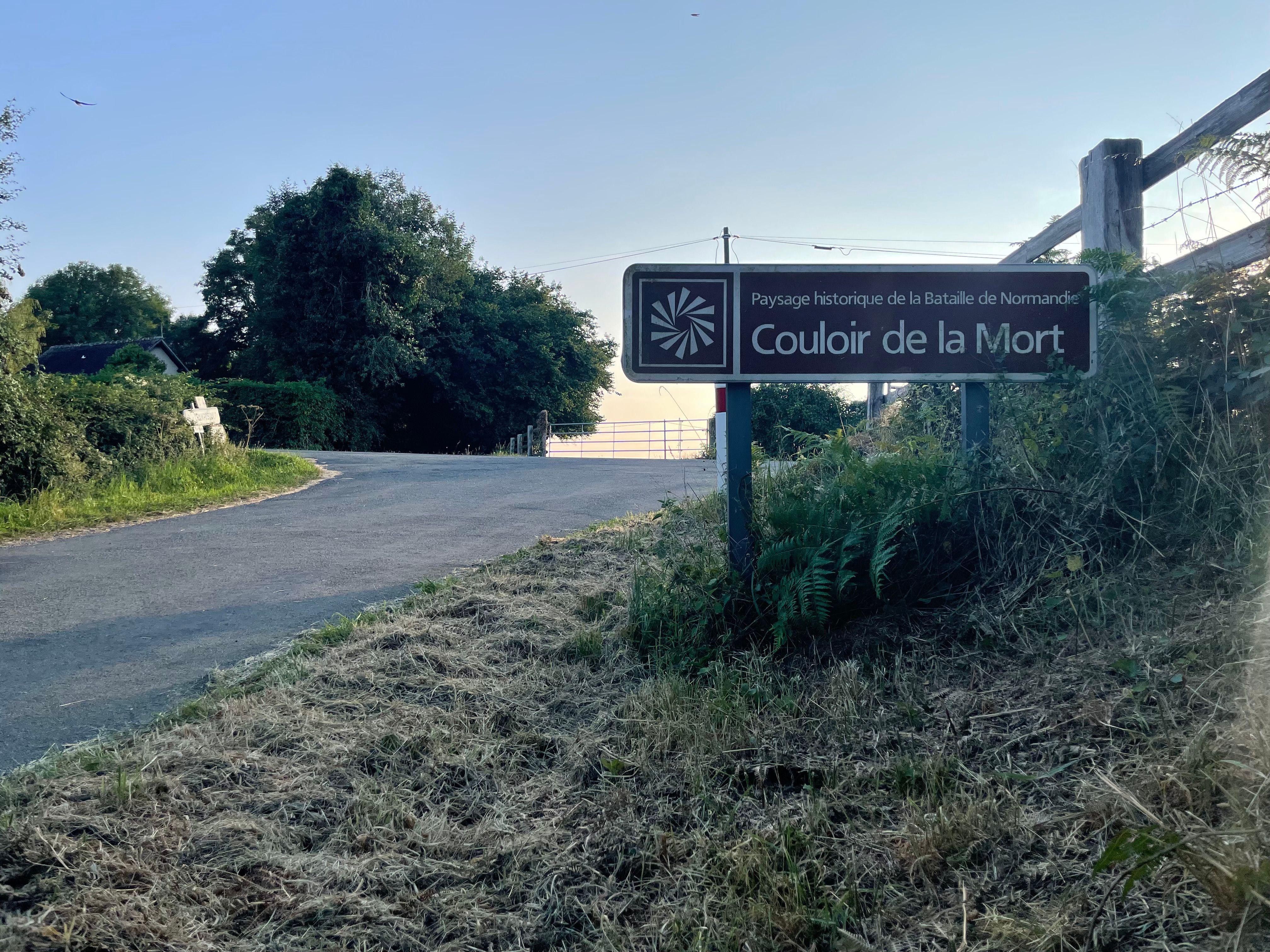 Panneau informatif et touristique: Paysage historique de la Bataille de Normandie – Couloir de la Mort. Au pied du panneau pousse une fougère.