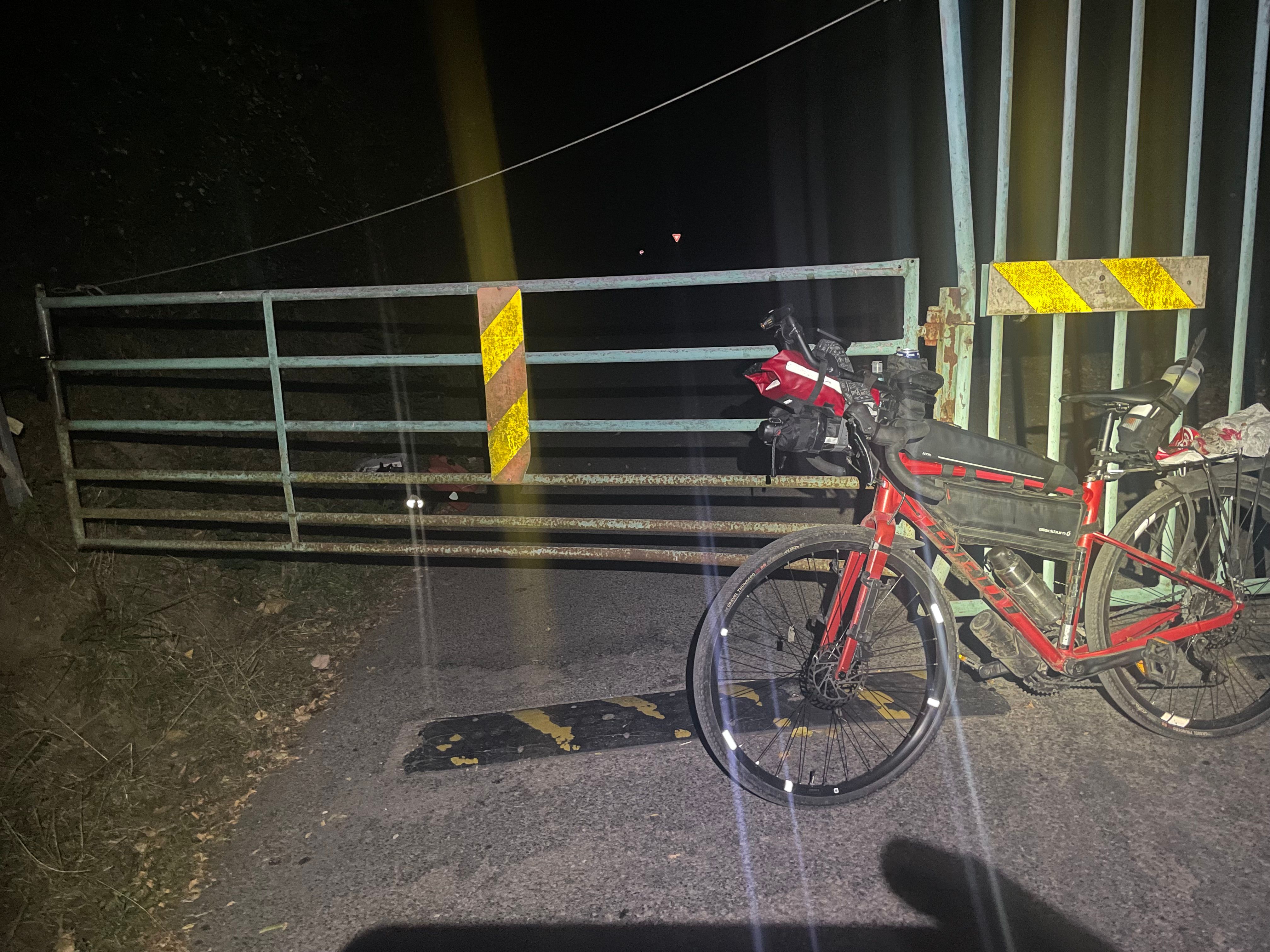 Photo de nuit. Le vélo est posé sur une barrière en 2 parties. Les principales sacoches sont déjà de l'autre coté de la barrière; À cause de l'objectif sale, les éléments réfléchissant de la barrière et du vélo dessinent des trainées lumineuses sur la photo